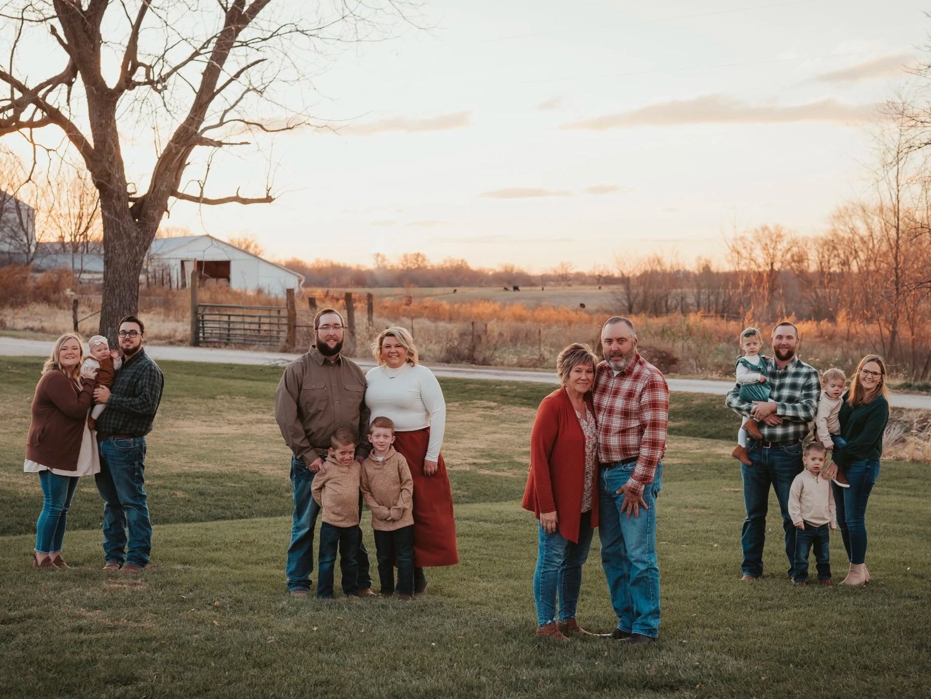 Family gathering in a grassy field at sunset, featuring multiple generations posing together. The scene includes adults and children dressed in casual fall attire, with a backdrop of trees and a rustic farm setting.