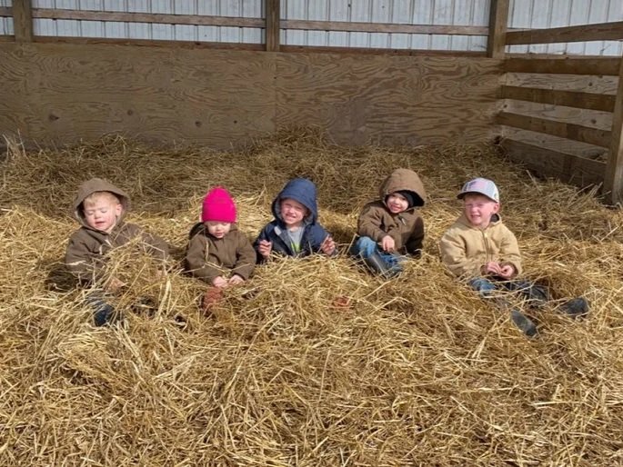 Five young children sitting in a barn filled with straw, wearing warm coats and hats, enjoying a playful moment together. The rustic setting highlights a cozy, farm-like atmosphere.